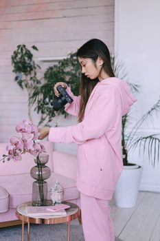 Asian woman in pink hoodie photographing orchids in a stylish indoor setting.