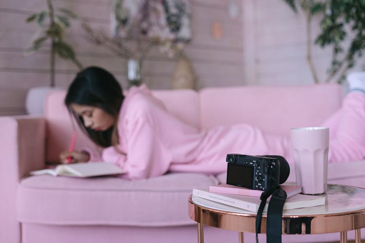 A Side Table With A Camera And A Cup Beside A Woman Taking Notes While Lying Down On A Couch