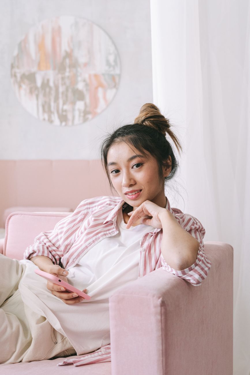 Young woman sitting on a pink sofa, using smartphone and smiling indoors.