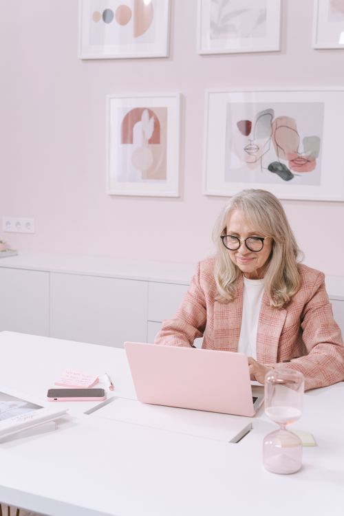 Free Senior woman in pink blazer using a laptop in a modern, artistic workspace. Stock Photo