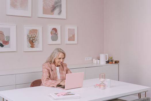 Elderly woman in pink blazer uses laptop in modern home office setting, smiling and engaged.