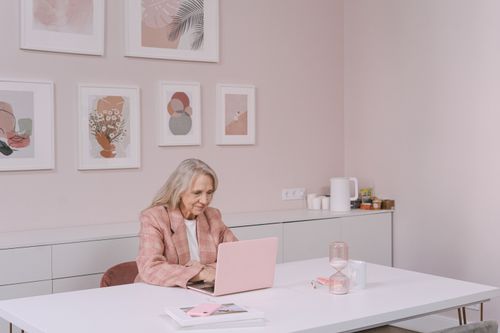 Free Elderly woman in pink blazer uses laptop in modern home office setting, smiling and engaged. Stock Photo