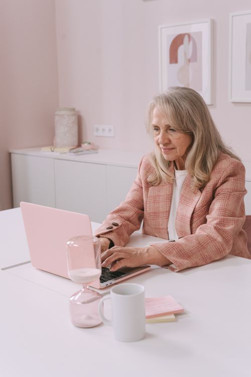 Free Elderly woman in pink blazer using laptop in a stylish pink office environment. Stock Photo