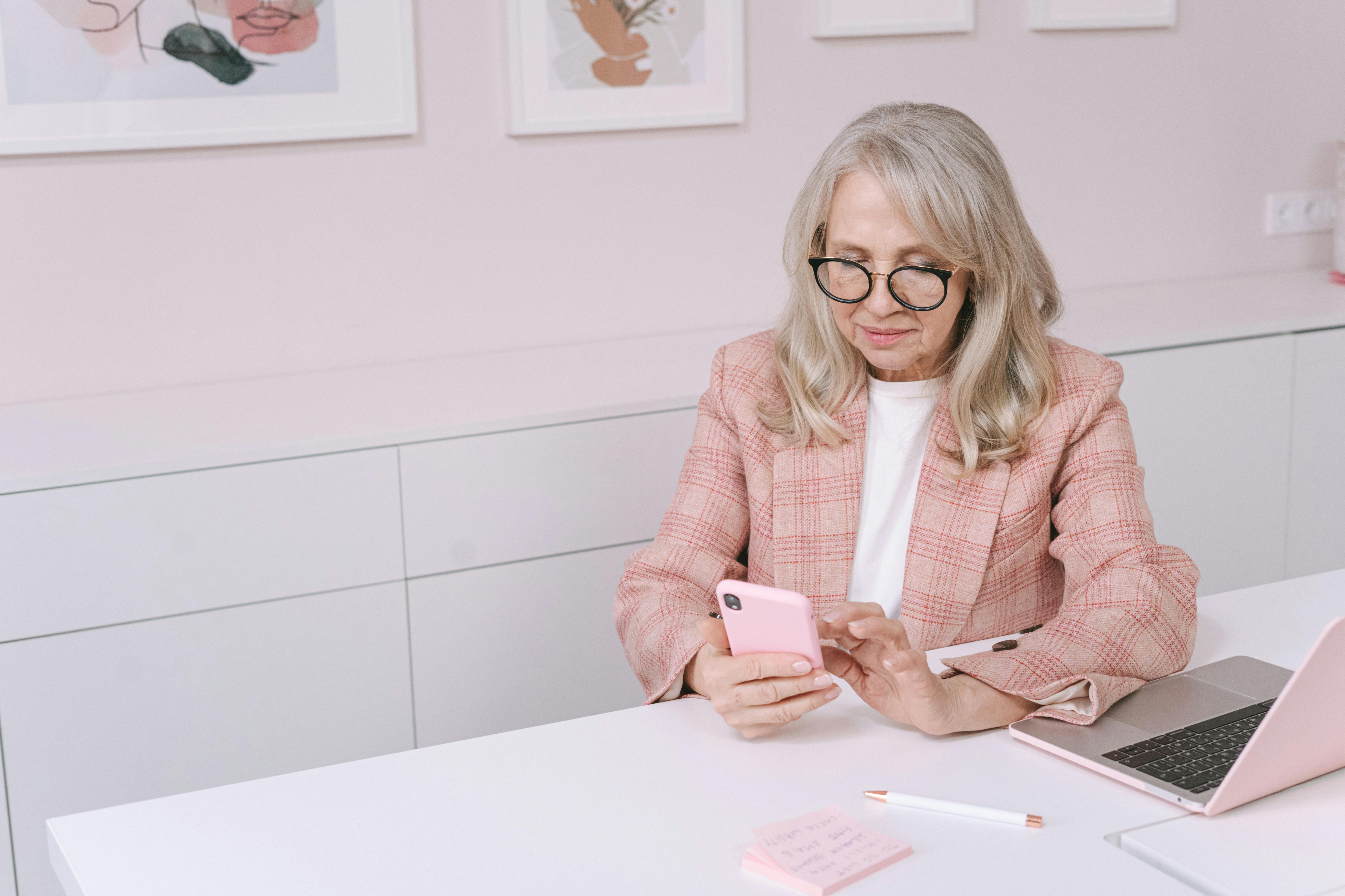 Elderly woman with eyeglasses using a smartphone at a desk, focusing on digital tasks in a stylish office.