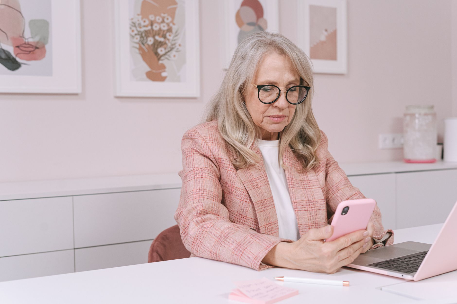 경남 어르신 스마트폰 무료 교육 2 Elderly woman in pink blazer using smartphone and laptop, working remotely from a stylish workspace.
