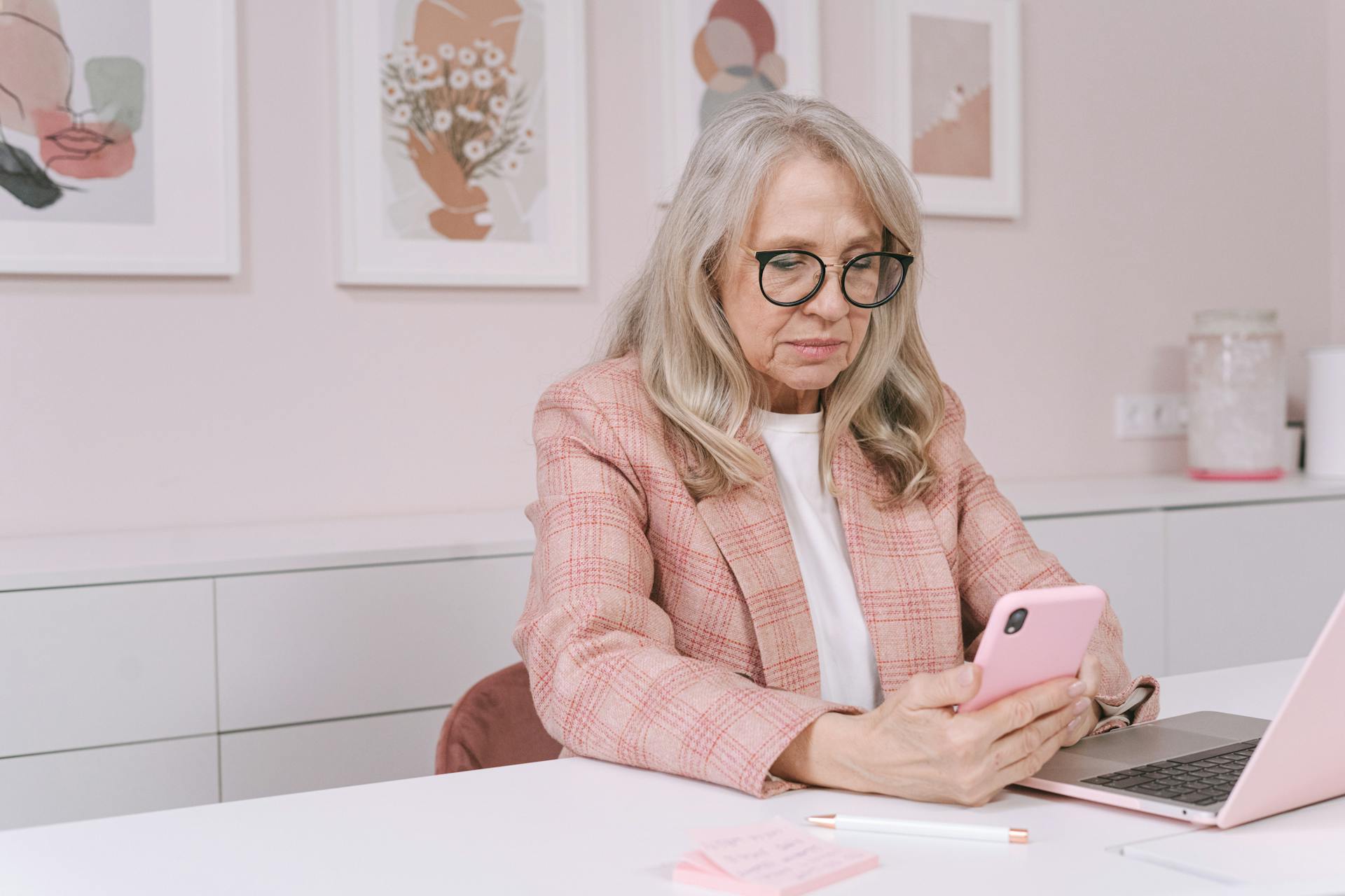 Elderly woman in pink blazer using smartphone and laptop, working remotely from a stylish workspace.