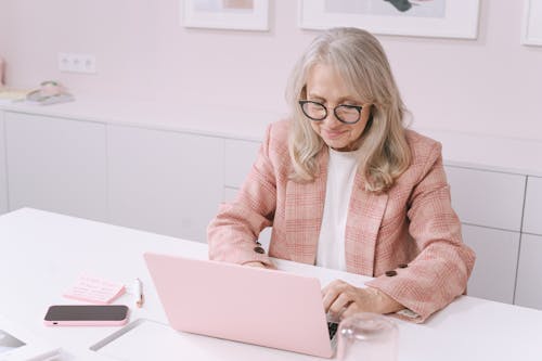 Elderly woman in pink blazer working on a laptop at a white table in a modern workspace.
