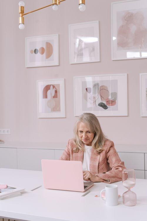 Free An elderly woman in a pink blazer works diligently on a pink laptop in a modern, art-filled office space. Stock Photo