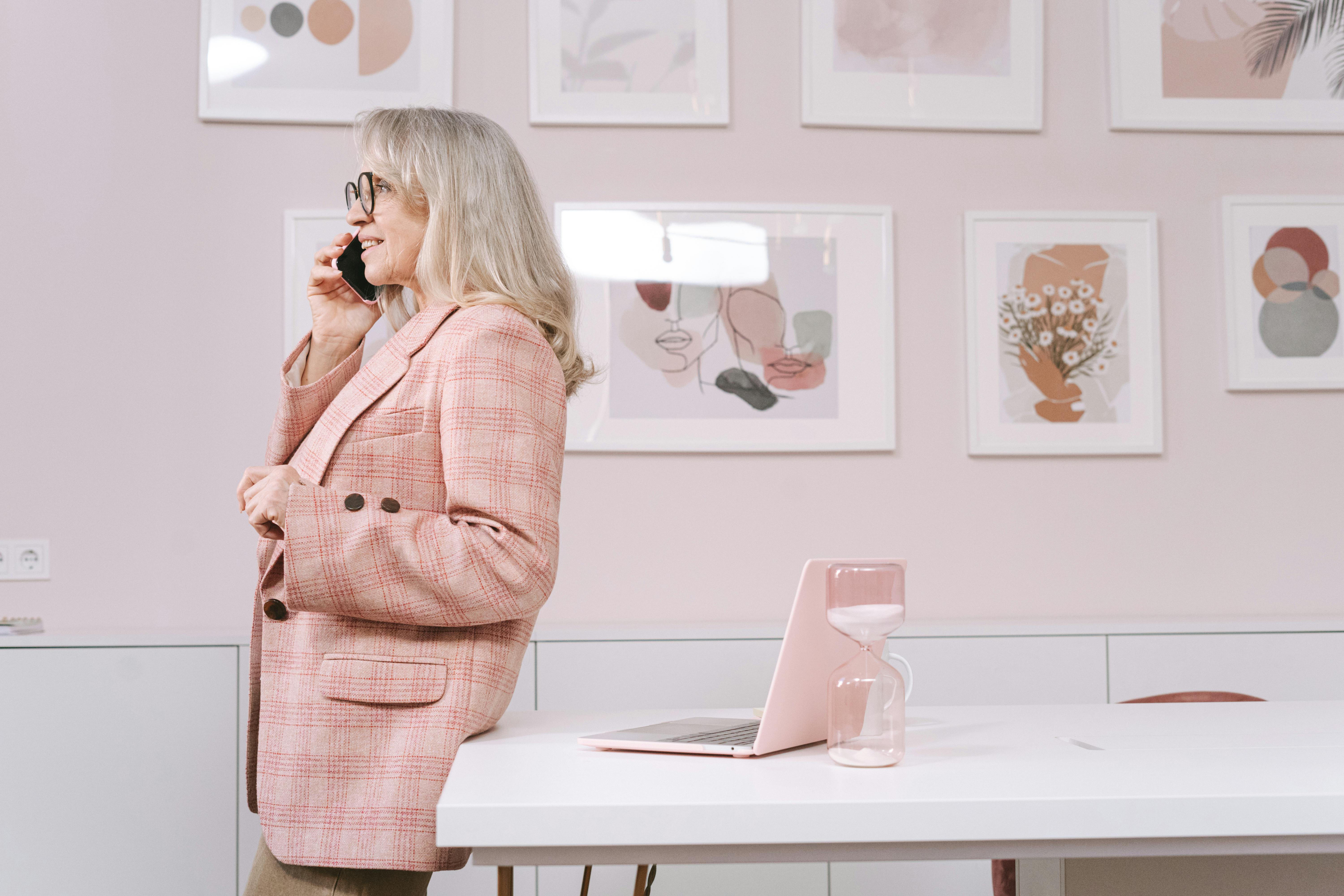 Senior woman in stylish blazer talks on smartphone in office, showcasing a modern professional environment with artwork.