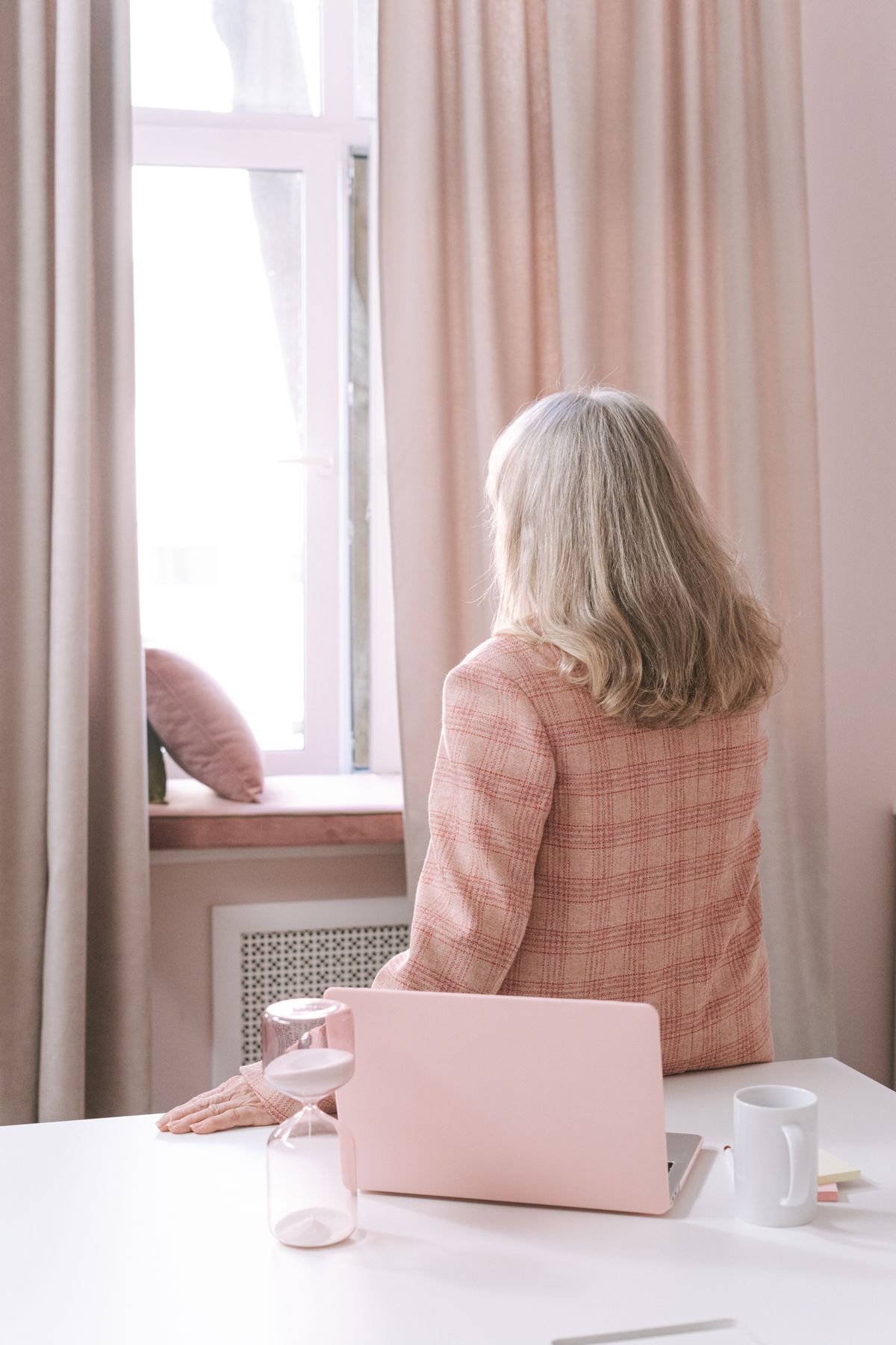A businesswoman with gray hair looks thoughtfully out a window in a stylish office with a laptop on the desk.