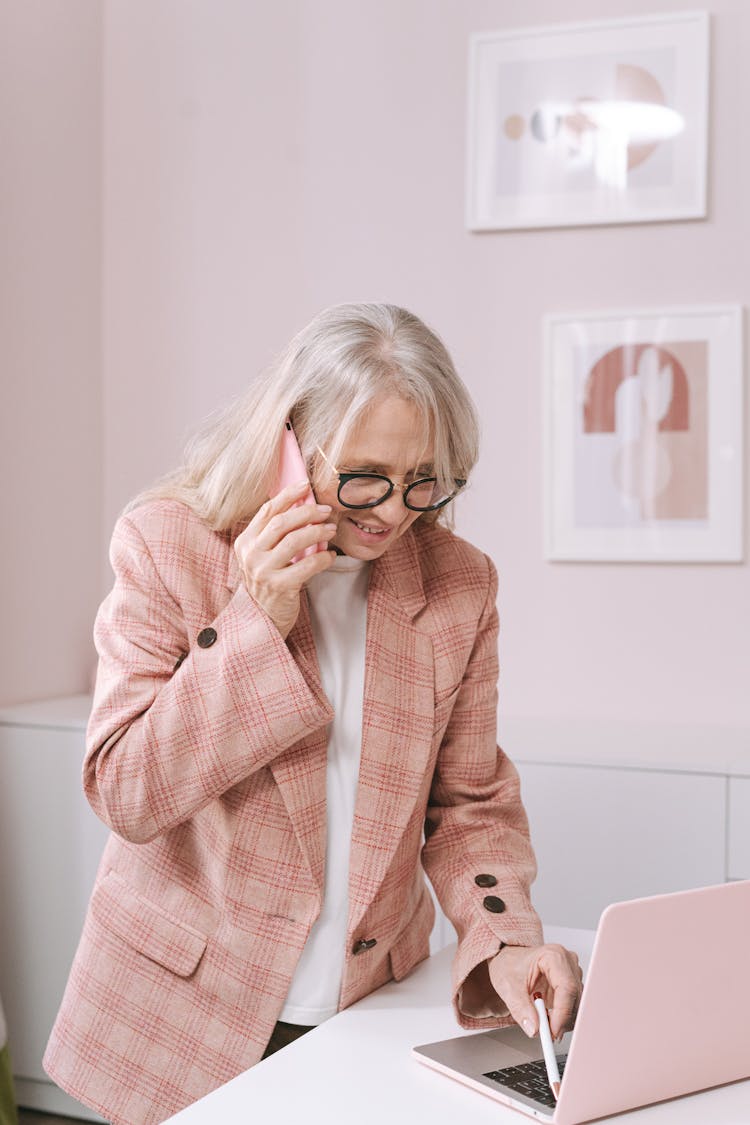 An Elderly Woman In A Pink Plaid Suit Using Her Phone While Holding A Pen To A Laptop