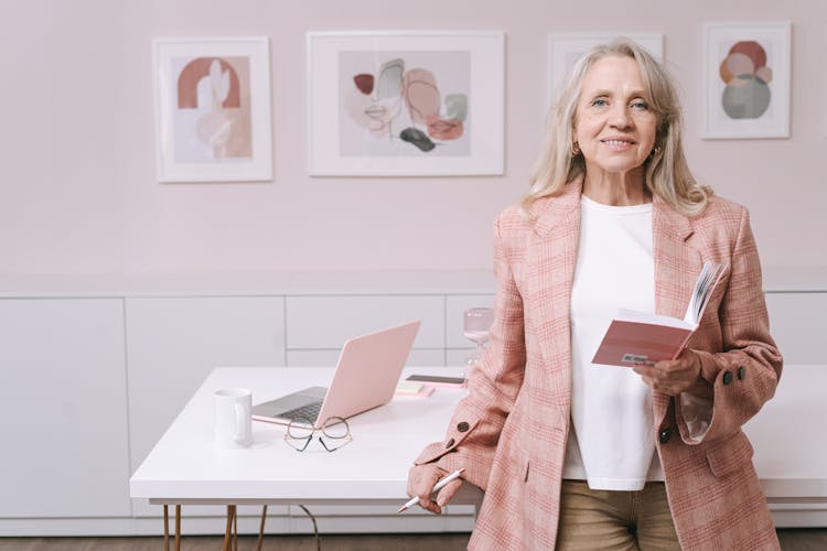 An Elderly Woman In Pink Blazer Smiling While Holding A Book