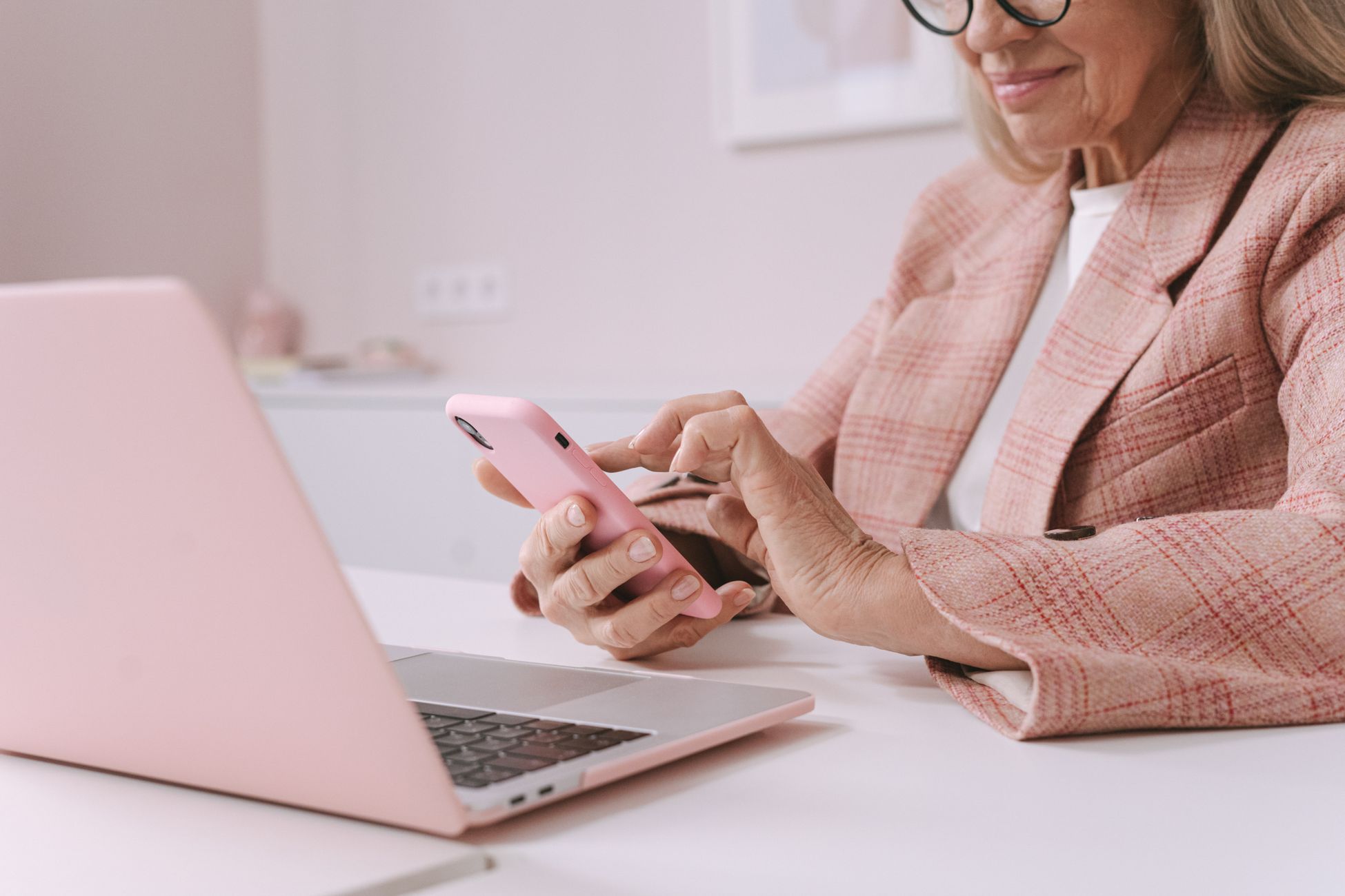 Elderly woman in pink plaid suit using a smartphone and laptop at a desk.