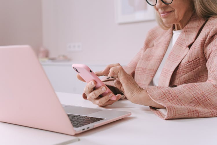 An Elderly Woman In Pink Plaid Suit Using A Smartphone In Front Of A Laptop