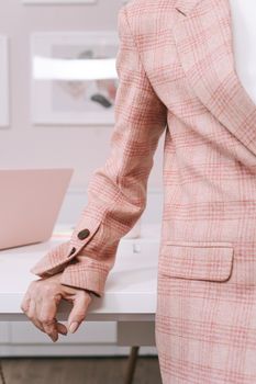 A person wearing a plaid blazer leans on a white desk in a stylish office with pink laptop.