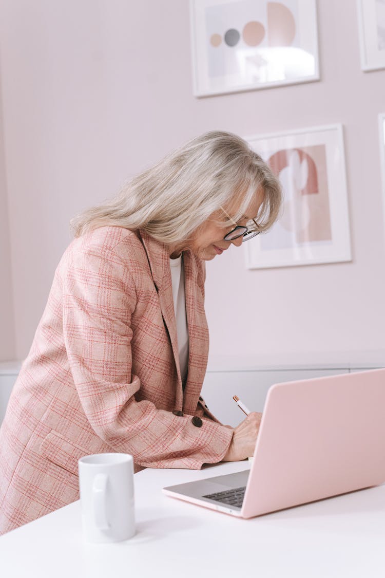 An Elderly Woman In Pink Blazer Writing On The Table