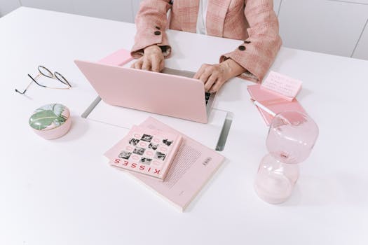 A stylish workspace with pink accessories including a laptop, books, and an hourglass on a white table.