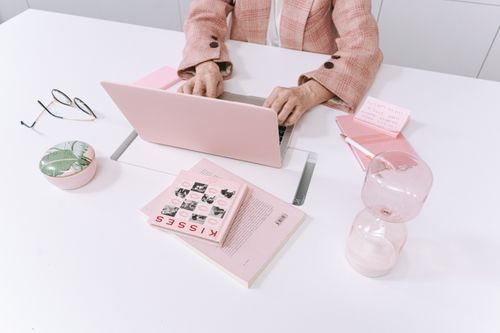 Free A stylish workspace with pink accessories including a laptop, books, and an hourglass on a white table. Stock Photo