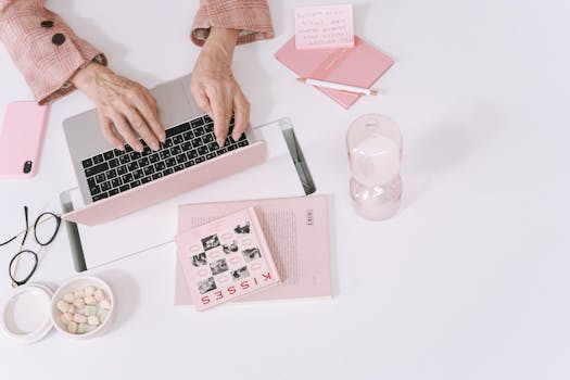 Chic flat lay of a feminine workspace featuring a laptop, pink stationery, and an hourglass.