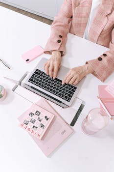 An adult typing on a laptop in a stylish pink-themed workspace with books and gadgets.