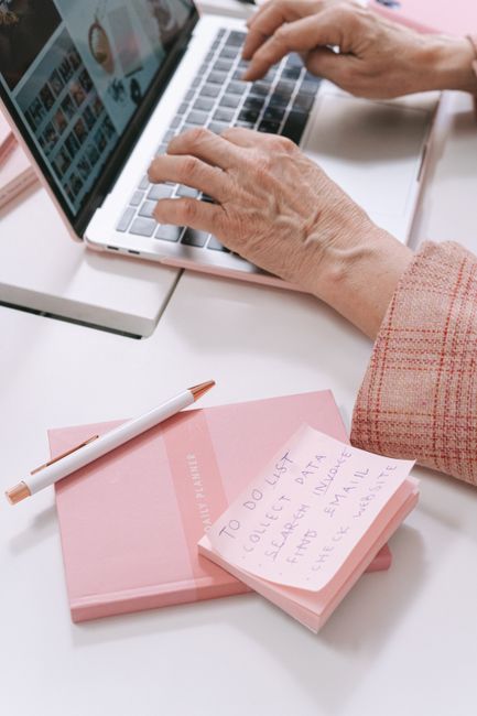 A person's hands typing on a laptop with a notebook, pen, and sticky notes on a white desk.