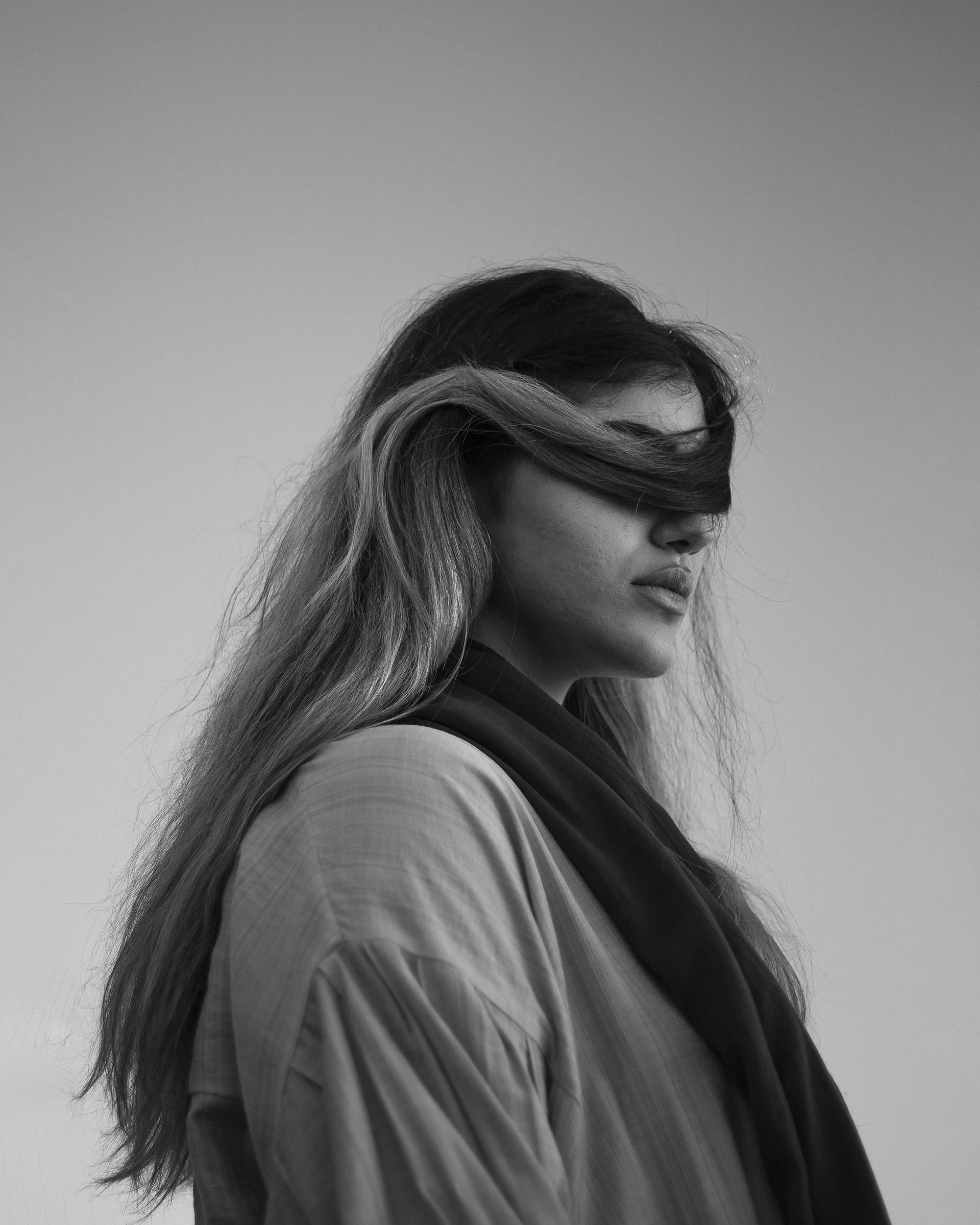 Black and white side view of anonymous female with hair covering eyes wearing outerwear and scarf standing against cloudless sky