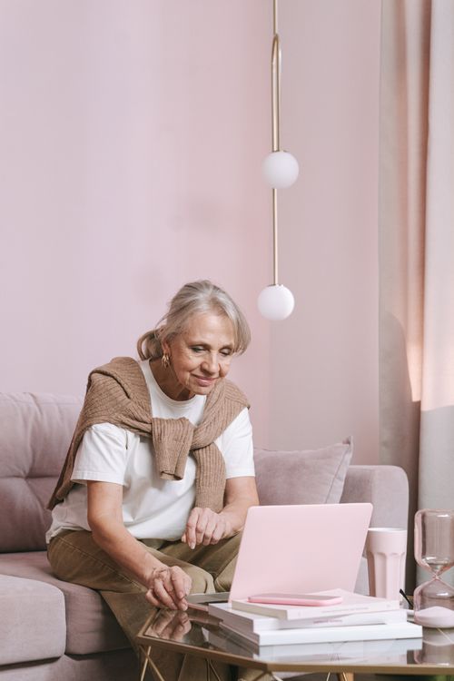 Free Elderly woman in sweater, using laptop on glass table in pink-themed room Stock Photo