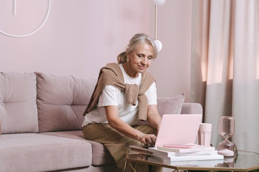 Elderly woman with gray hair smiling while working on a laptop at home, enjoying remote work.