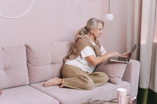 Free Elderly woman sitting comfortably using a laptop on a pink couch at home. Stock Photo