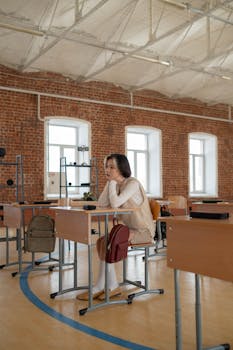 Young woman sitting thoughtfully at a desk in a spacious classroom with brick walls.