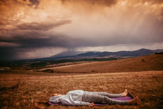 Person meditating on a yoga mat in a serene countryside setting with moody clouds.