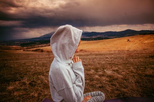Person meditating in hoodie outdoors with mountains and cloudy sky in view.
