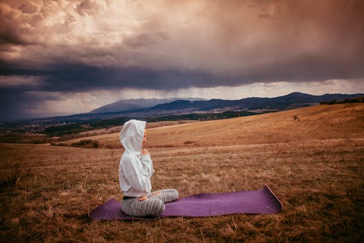 Woman meditating on a yoga mat amidst a serene countryside landscape with cloudy skies.