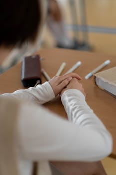 A student sitting at a desk with hands folded, pencils, and books in a classroom setting.