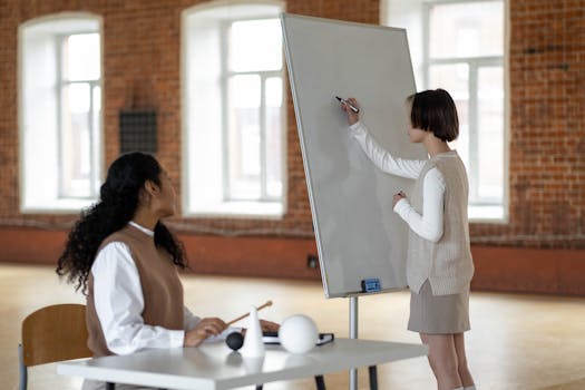 A student writes on a whiteboard watched by a teacher in a classroom setting.