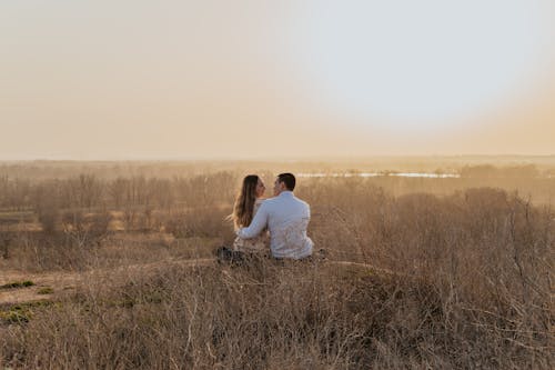 Back view of tender loving couple sitting on hill and hugging while enjoying sunset in nature