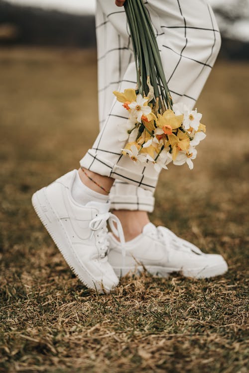 Side view of crop anonymous female in checkered pants and trendy footwear standing on lawn with blossoming daffodils in daytime