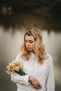 Young tender female in white shirt with blooming daffodils and wavy blond hair against pond in daylight