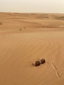 Stylish sunglasses resting on golden desert dunes under a hazy sky, showcasing serene and vast landscapes.