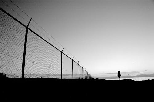 Black and white silhouette of anonymous person standing alone in field near net fence