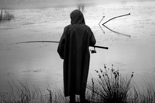 Black and white of faceless fisherman with rod standing on shore of pond and fishing