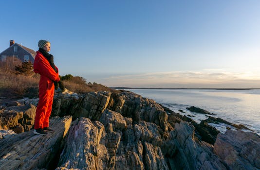 A woman in a red outfit stands on rocky cliffs, enjoying the serene ocean sunrise view on a calm morning.