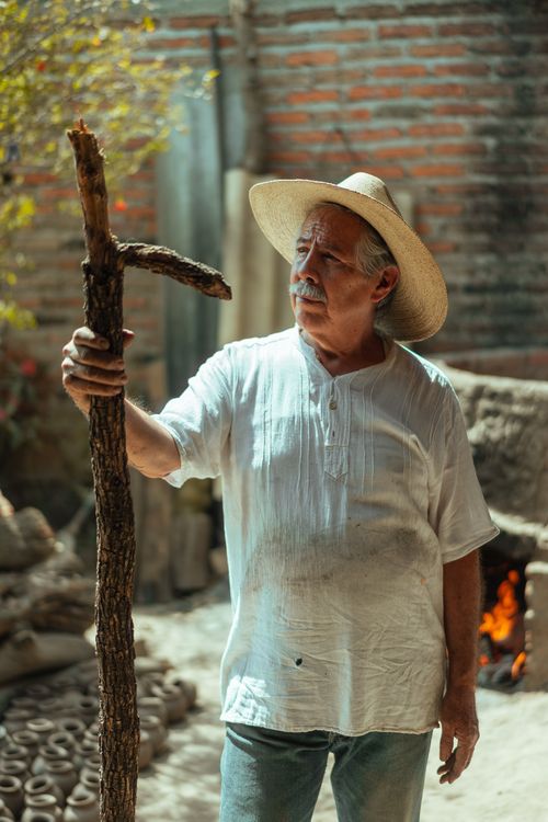 Free Elderly man in traditional attire holding a wooden stick with a kiln in the background, Tonalá, Mexico. Stock Photo