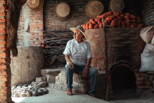 Free A senior artisan in a straw hat sits surrounded by earthenware pottery in a rustic workshop. Stock Photo