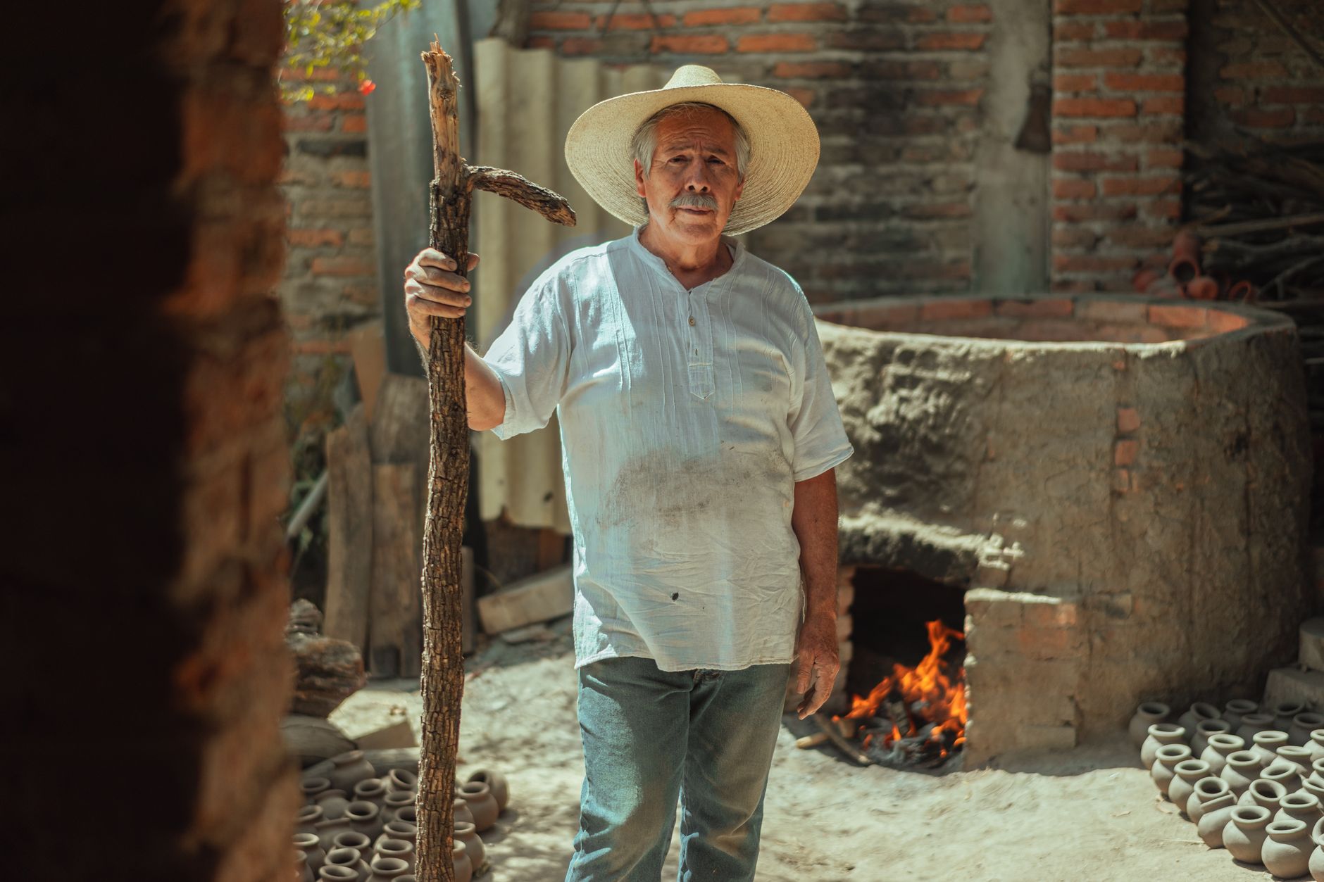 Senior potter stands outdoors with kiln, clay pots, and tools highlighting traditional craftsmanship.