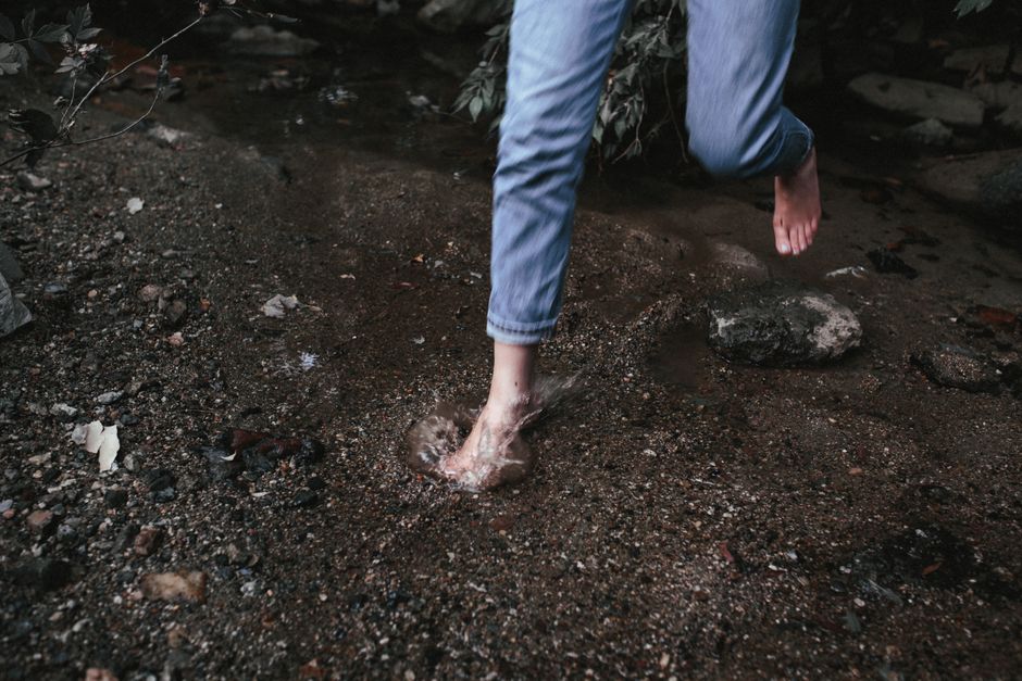 A person wearing blue jeans jumps barefoot in a rocky stream.