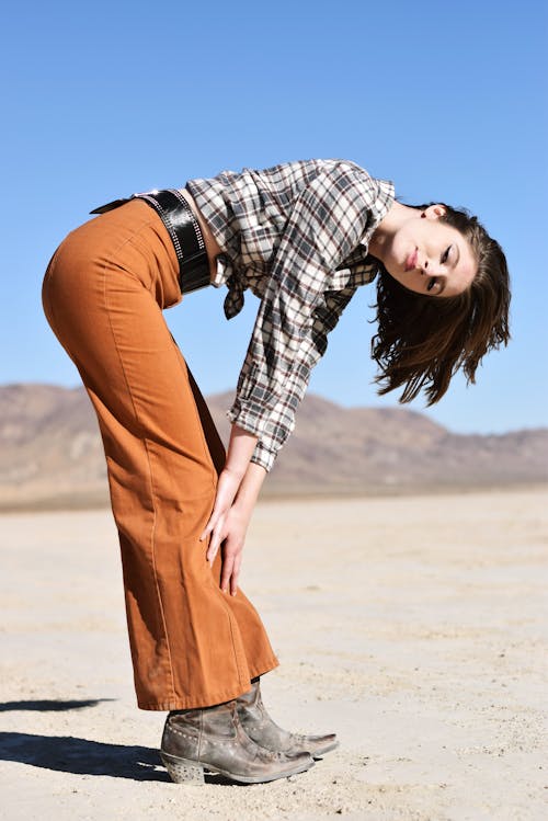 Side view of stylish young woman in checkered shirt and pants touching legs while looking at camera under blue sky