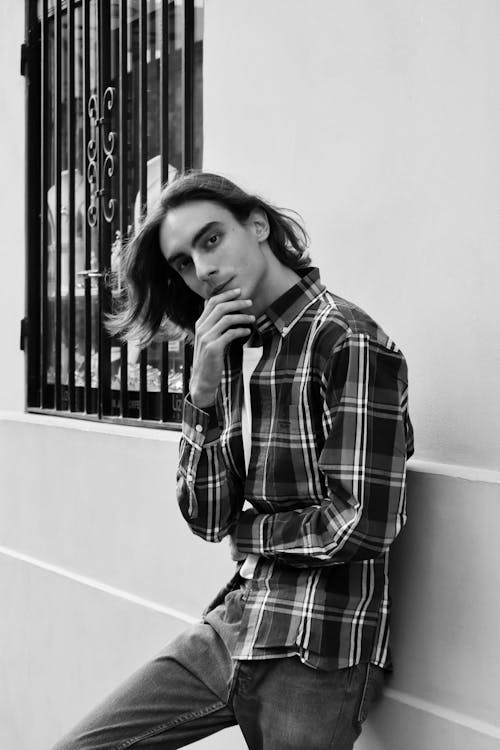 A Grayscale Photo of a Man in Plaid Long Sleeves Leaning on the Wall