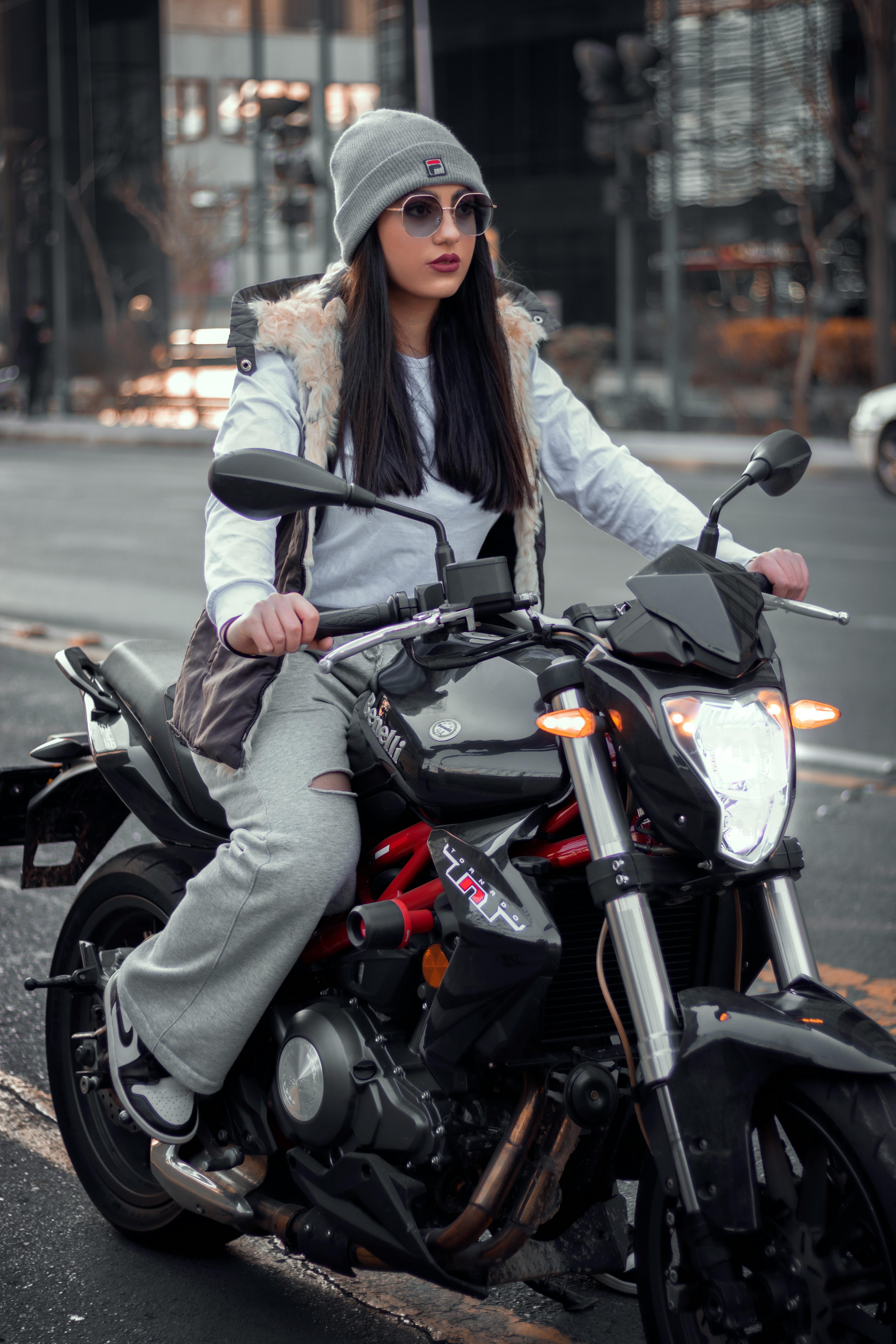 A fashionable woman on a motorcycle in the city, wearing casual gear and sunglasses.