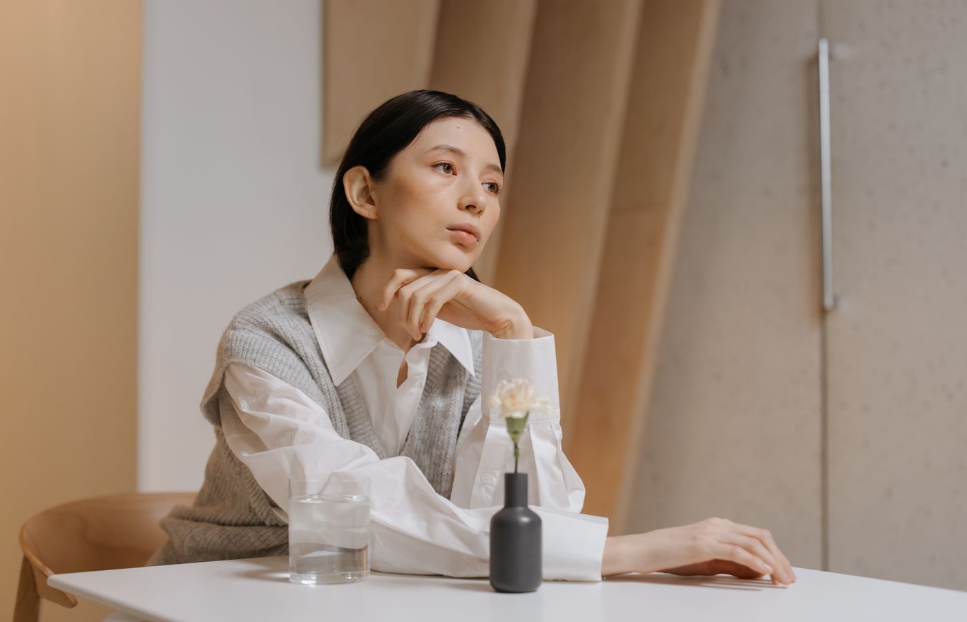 Thoughtful woman in white blouse and vest sitting at a table indoors, lost in deep thought.